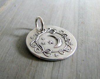 May include: A silver charm pendant featuring a crescent moon and stars surrounded by a decorative scroll design.