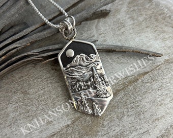 Könnte beinhalten: Eine silberne Halskette mit einem Anhänger, der eine detaillierte Darstellung einer Bergkette mit einem Vollmond am Himmel zeigt. Der Anhänger ist schildförmig und hat eine strukturierte, fast holzartige Oberfläche.