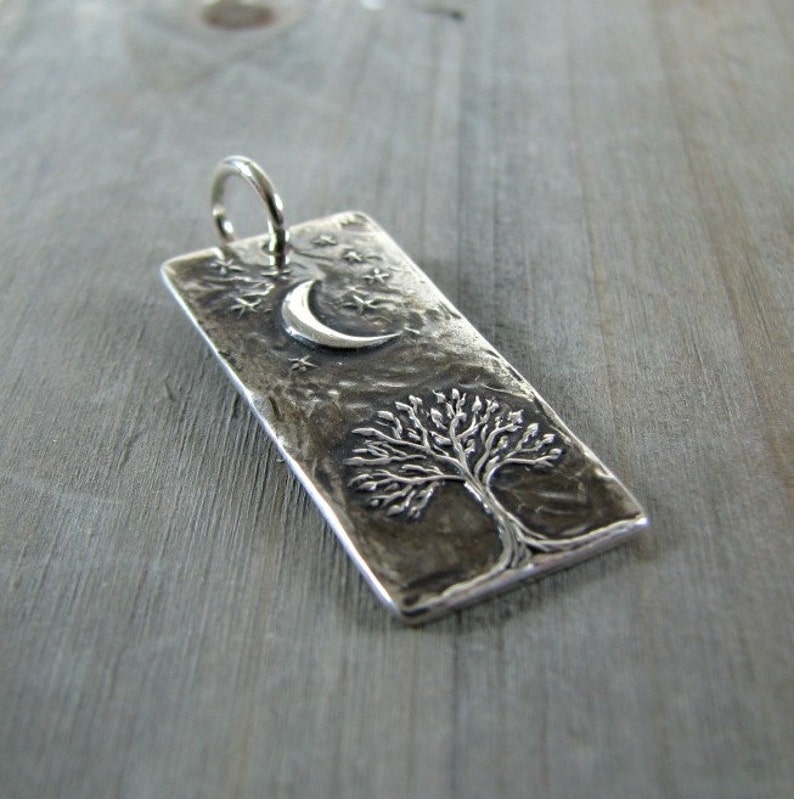 Puede incluir: Un colgante de plata con una superficie texturizada que presenta un &aacute;rbol y una luna creciente con estrellas.