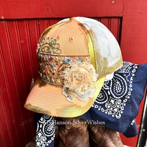 May include: A peach-colored trucker hat with a gold brim and a white mesh back. The hat is embellished with floral embroidery, a large beige fabric flower, and gold trim. A navy blue paisley bandana is draped beneath the hat.