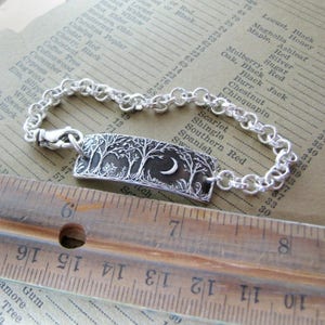 May include: A silver bracelet with a charm featuring a crescent moon and trees. The charm is rectangular and has a textured surface. The bracelet is made of a silver chain with a lobster clasp.