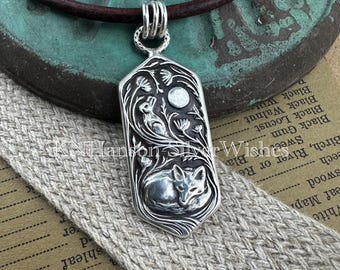 Puede incluir: Colgante de plata con un diseño en relieve detallado de un zorro durmiente, un conejo, una luna y elementos florales. El colgante es rectangular con bordes redondeados y cuelga de un cordón de cuero marrón. El colgante mide aproximadamente 3,8 cm de largo.