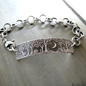May include: A silver bracelet with a charm featuring a crescent moon and a detailed silhouette of trees. The bracelet has a chain link design.