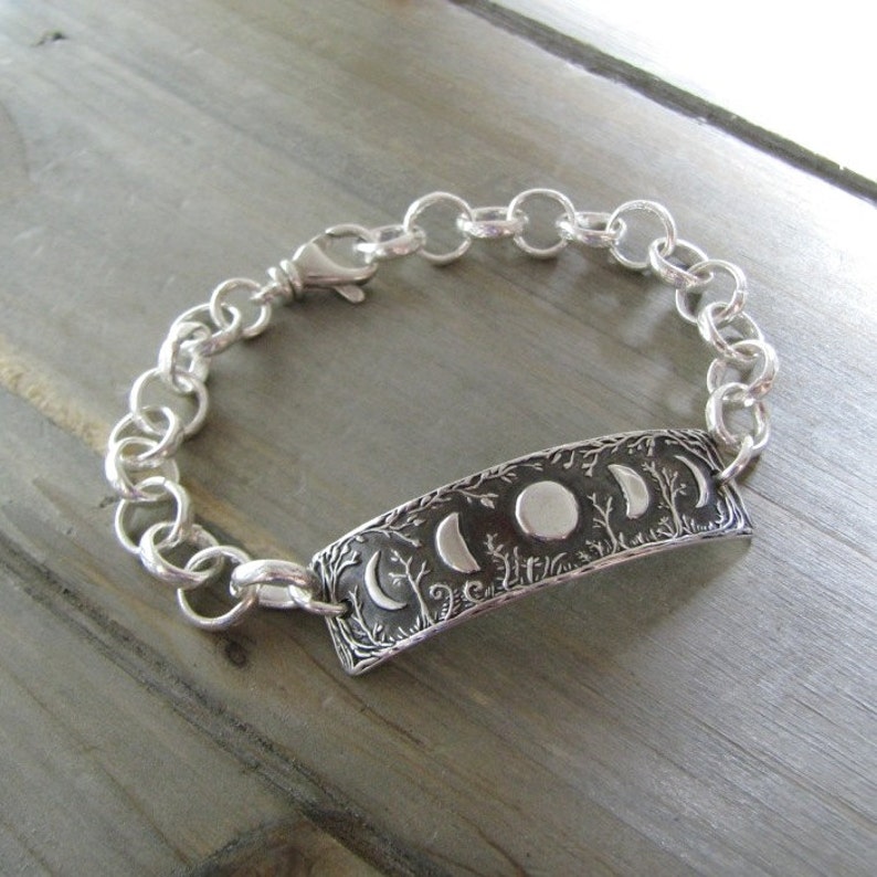 Puede incluir: Una pulsera de plata con un dije que presenta un dise&ntilde;o detallado de una luna con fases y &aacute;rboles. La pulsera est&aacute; hecha de una cadena de plata.
