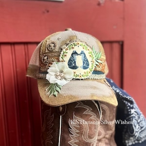 May include: A beige and gold baseball cap with a decorative floral design and a patch featuring two blue foxes. The cap is embellished with a white flower and green leaves. The brim is a light beige colour.