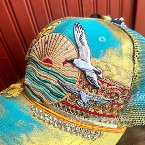 Peut inclure: Casquette de baseball brodée avec un motif turquoise et or. Représente un lever de soleil sur l'océan et deux mouettes. La casquette a un dos en filet et est ornée de bordures dorées et de strass.