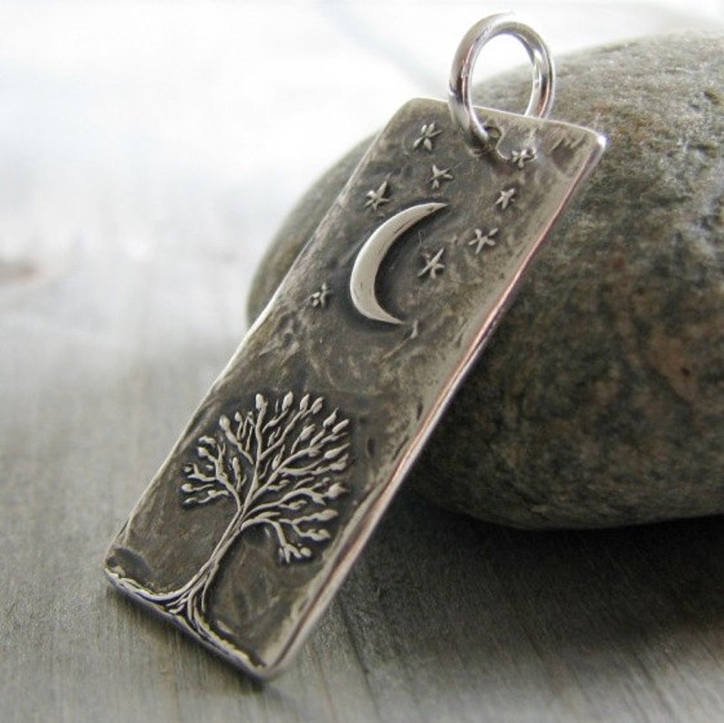 Puede incluir: Colgante de plata con una superficie texturizada que presenta un &aacute;rbol y una luna creciente con estrellas.