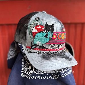 May include: A gray trucker hat with a distressed brim and a black mesh back. The hat features an embroidered owl design with a red, turquoise, and black color scheme. The brim is decorated with rhinestones and a red trim.