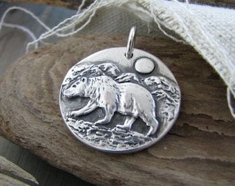 Puede incluir: Un colgante de plata con un oso caminando frente a una cordillera con luna llena en el fondo.