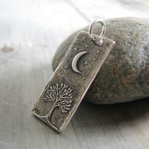 Puede incluir: Un colgante de plata con un dise&ntilde;o de &aacute;rbol texturizado, una luna creciente y estrellas. El colgante es rectangular y tiene un acabado martillado.