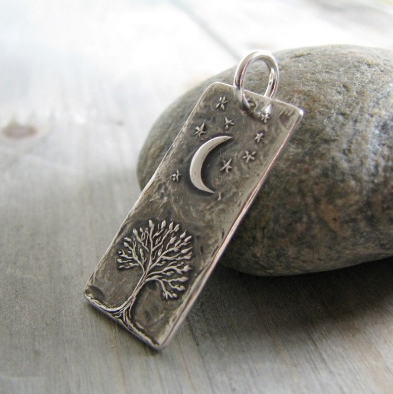 Puede incluir: Un colgante de plata con un dise&ntilde;o de &aacute;rbol texturizado, una luna creciente y estrellas. El colgante es rectangular y tiene un acabado martillado.