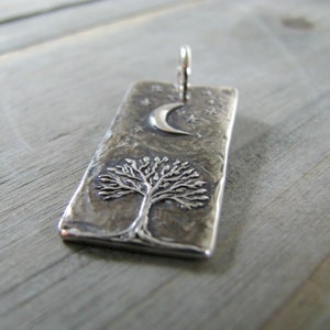 Puede incluir: Un colgante de plata con una superficie texturizada que presenta un &aacute;rbol, una luna creciente y estrellas. El colgante es rectangular y tiene un bucle en la parte superior para sujetar una cadena.