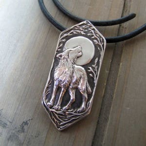 May include: Silver pendant necklace featuring a wolf howling at the moon. The pendant is detailed with a textured background and a black leather cord.