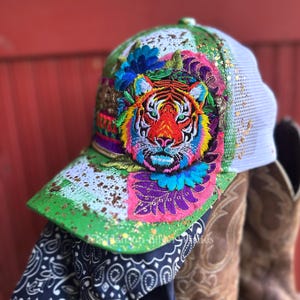 Könnte beinhalten: Eine Baseballkappe mit einem lebendig bestickten Tigergesicht, umgeben von bunten Blumen- und Blattmustern. Die Kappe hat einen grünen Schirm mit goldenen Farbspritzern, ein weißes Netzgewebe auf der Rückseite und den Schriftzug KNHanson-SilverWishes.
