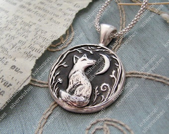 Puede incluir: Un collar de plata con un colgante redondo que presenta una representación detallada de un zorro sentado bajo una luna creciente. El colgante está rodeado de intrincados diseños florales.