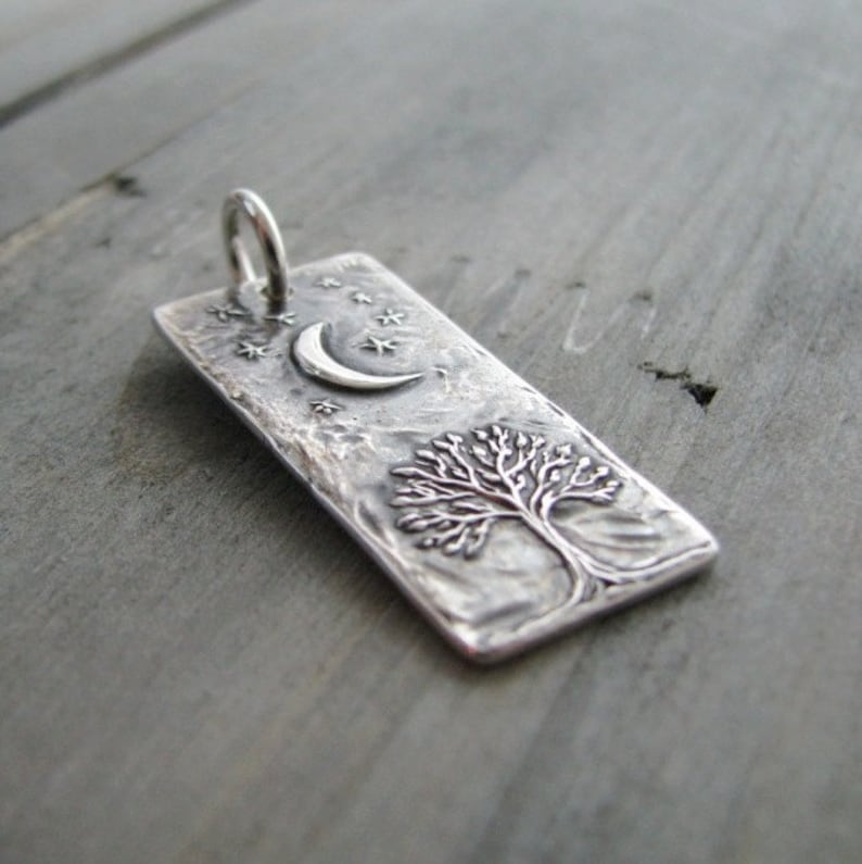 Puede incluir: Un colgante de plata con una superficie texturizada que presenta un &aacute;rbol y una luna creciente con estrellas.