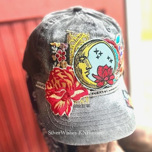 May include: A charcoal gray distressed baseball cap featuring a large embroidered patch of a yellow crescent moon and a red lotus flower. The cap is decorated with additional floral embroidery, gold patterned fabric accents, and a thin rhinestone trim.