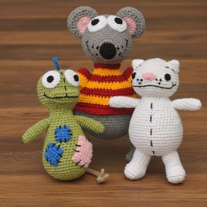 May include: Three crocheted soft toys: a grey mouse in a red and yellow striped top, a green creature with blue and pink patches, and a white cat with a pink nose. They are displayed on a wooden surface.