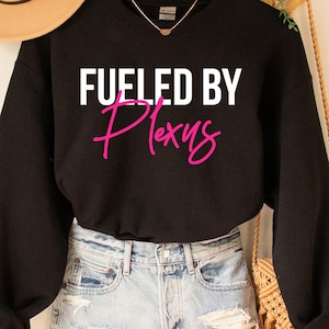 Può includere: Una felpa nera con la scritta "FUELED BY Plexus" in lettere bianche e rosa. La felpa è abbinata a pantaloncini di jeans e un cappello beige. Sono visibili anche una gruccia di legno e una borsa in macramè.