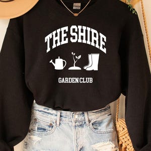 May include: A black sweatshirt with the text "THE SHIRE GARDEN CLUB" in white. The design includes a watering can, a plant, and a boot. A tan hat and denim shorts are also visible.
