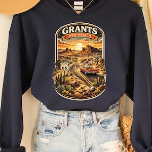 May include: A navy blue sweatshirt with a graphic featuring a desert landscape, a vintage car, and the text "Grants New Mexico 66". The sweatshirt is paired with denim shorts and a tan hat.