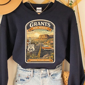 May include: A navy blue sweatshirt with a Route 66 graphic featuring the text "Grants, New Mexico." The graphic depicts a road scene with a vintage car, mountains, and a church. A tan hat and denim shorts are also visible.