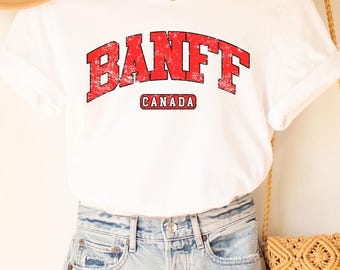 以下が含まれることがあります： 赤色の「BANFF」の文字と「CANADA」がプリントされた白いTシャツ。ライトウォッシュデニムのショートパンツ、タンカラーの帽子、フリンジ付きの織りクロスボディバッグと組み合わせています。