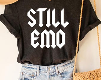 May include: A dark gray t-shirt with the text "STILL EMO" in white, paired with denim shorts, a tan hat, and a woven crossbody bag. The outfit is displayed on a wooden hanger against a neutral background.