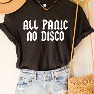 May include: A black t-shirt with the text "All Panic No Disco" in white letters. The shirt is styled with a brown hat, denim shorts, and a crocheted bag.