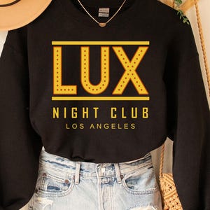 May include: Black sweatshirt with the text "LUX NIGHT CLUB LOS ANGELES" in yellow. The sweatshirt is hanging on a wooden hanger. A tan hat and denim shorts are also in the image.