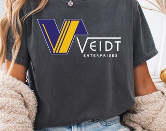Könnte beinhalten: Dunkelgraues T-Shirt mit dem Logo "VEIDT ENTERPRISES" in Weiß, Gelb und Blau. Das T-Shirt hat einen Rundhalsausschnitt und kurze Ärmel. Das T-Shirt wird mit Jeansshorts getragen.
