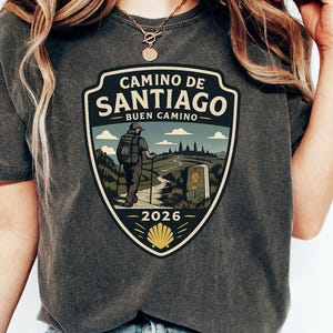 May include: A charcoal gray short-sleeve t-shirt featuring a graphic shield emblem with the text Camino de Santiago, Buen Camino, and 2026. The illustration depicts a hiker with a backpack and walking poles on a trail, a milestone, and a yellow shell.
