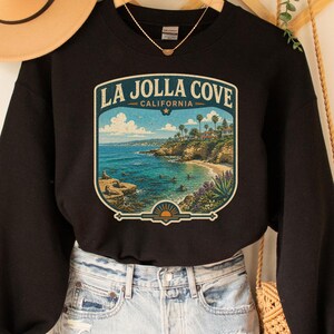 May include: Black sweatshirt with a graphic of La Jolla Cove, California. The design features a coastal scene with blue water, a sandy beach, and palm trees. The sweatshirt is paired with denim shorts and a tan hat.