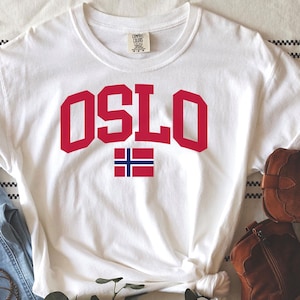 May include: A white t-shirt with the word "OSLO" in red block letters and a Norwegian flag graphic. The shirt is styled with light blue jeans and brown leather cowboy boots. A set of bracelets and a sprig of greenery complete the look.