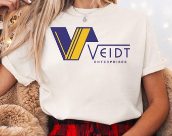 Könnte beinhalten: Cremefarbenes T-Shirt mit dem Text "VEIDT ENTERPRISES" in Marineblau und Gelb. Das Shirt hat ein stilisiertes Logo. Die Person trägt ein Armband und sitzt vor einem Weihnachtsbaum.