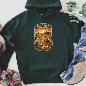 May include: Dark green hoodie with a Grants, New Mexico graphic. The graphic features a desert landscape with a road, a vintage car, and the text "Grants New Mexico" in an oval shape. The hoodie is paired with jeans, boots, and a plaid scarf.