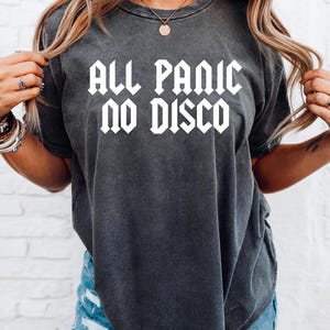 May include: Dark gray t-shirt with the text "ALL PANIC NO DISCO" in white, gothic-style lettering. The shirt has short sleeves and a slightly faded appearance. The model is wearing blue denim shorts.