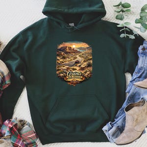 May include: Dark green hoodie featuring a desert landscape graphic with the text "Grants New Mexico." The image includes blue jeans, tan boots, and a plaid scarf. The outfit suggests a relaxed, casual style.