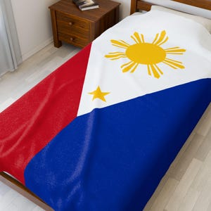 Könnte beinhalten: Eine Bettdecke mit der Flagge der Philippinen. Das Design umfasst ein weißes Dreieck mit einer gelben Sonne und drei Sternen sowie rote und blaue Felder. Die Bettdecke liegt auf einem Bett, im Hintergrund ist ein Nachttisch aus Holz zu sehen.