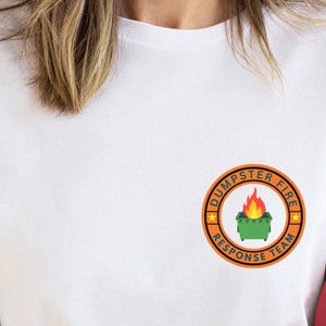May include: A white short-sleeved t-shirt featuring a circular graphic on the left chest. The graphic has an orange border with the text "DUMPSTER FIRE RESPONSE TEAM" and depicts a green dumpster with orange and yellow flames rising from it.