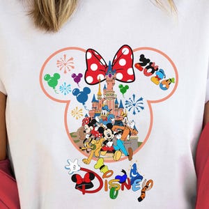 May include: White t-shirt featuring a colorful Disney graphic. The design includes a red polka-dot bow, a castle, and various Disney characters. The word "Disney" is also displayed in multiple colors.
