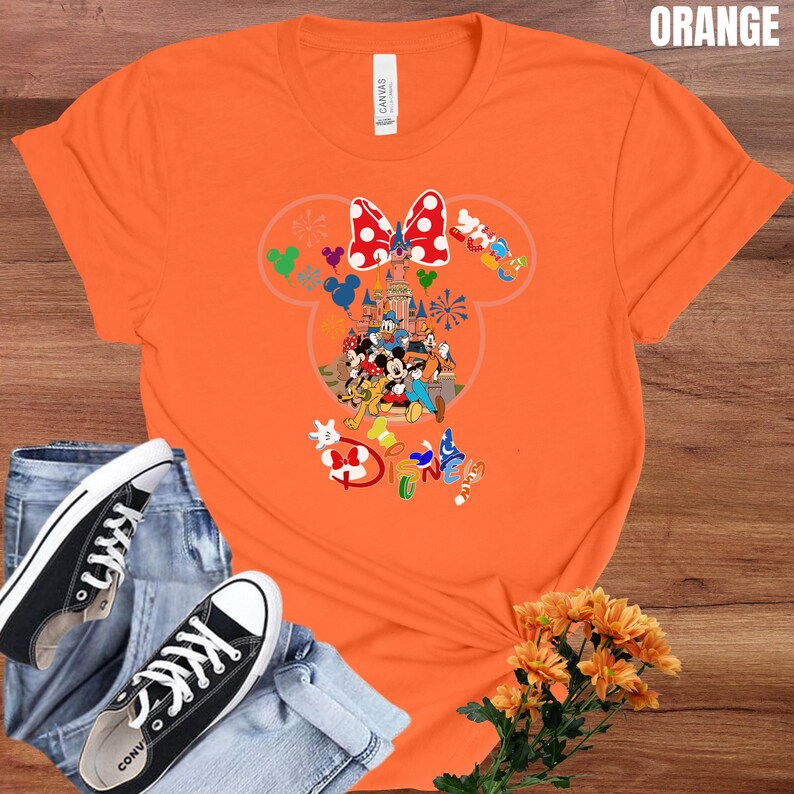 May include: Orange t-shirt featuring a Minnie Mouse design with a red and white polka dot bow, a castle, and various Disney characters. The word "Disney" is also visible. The shirt is paired with blue jeans and black sneakers.