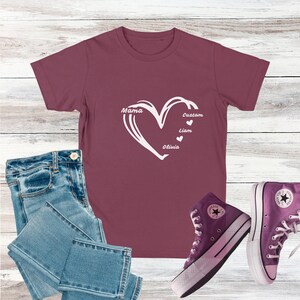 May include: A maroon t-shirt with a white heart design, featuring the words "Mama", "Custom", "Liam", and "Olivia". The shirt is paired with blue jeans and purple high-top sneakers.