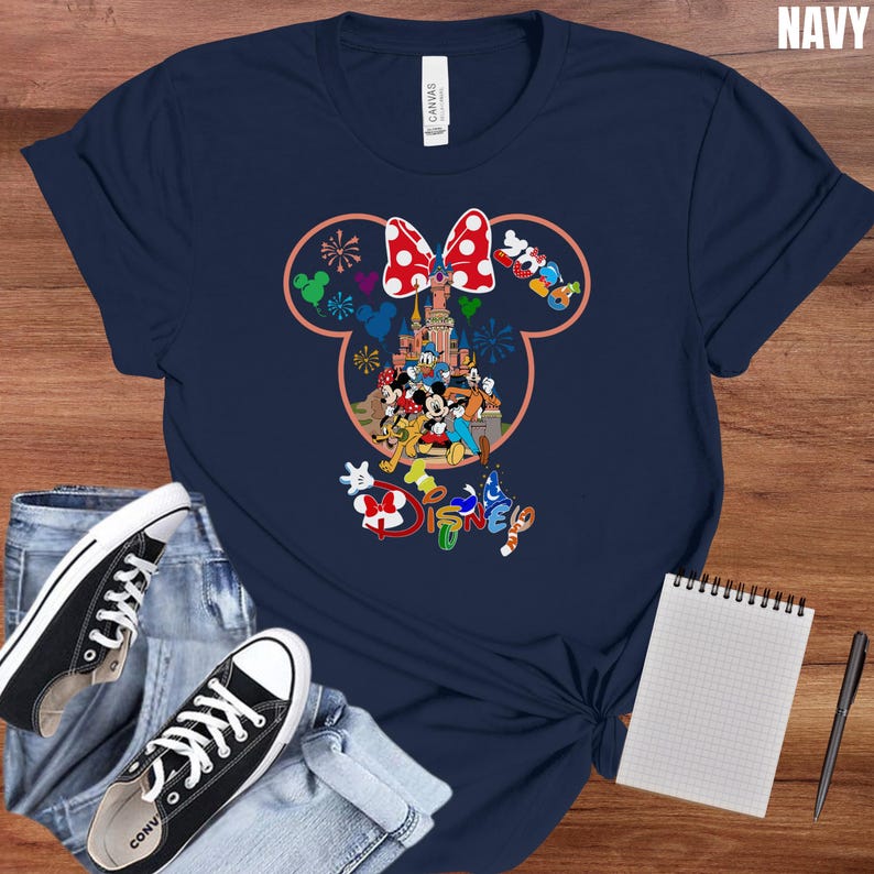 May include: Navy blue t-shirt featuring a colorful Disney design. The graphic includes a castle, Mickey Mouse, Minnie Mouse, and other characters. The word "DISNEY" is also displayed. The shirt is paired with black sneakers and blue jeans.