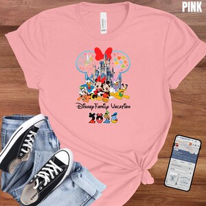 May include: Pink t-shirt featuring a Disney Family Vacation design with Mickey and Minnie Mouse, Donald Duck, Daisy Duck, and Goofy. The design includes a castle, a bow, and the year 2026. Black canvas sneakers and blue jeans are also in the image.