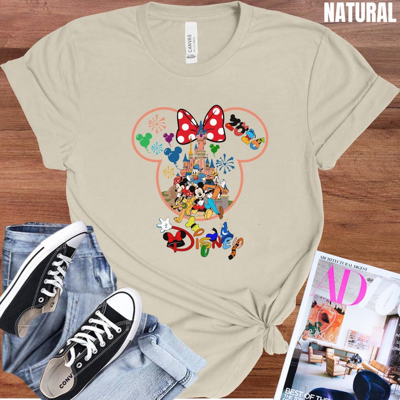 May include: A beige t-shirt featuring a colorful Disney graphic. The design includes a red bow with white polka dots, a castle, and various Disney characters. The word "Disney" is also displayed in colorful letters.