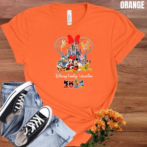 May include: Orange t-shirt featuring a Disney Family Vacation design with Mickey and Minnie Mouse, Donald Duck, and other characters. The design includes a castle and the year 2026. The shirt is paired with blue jeans and black sneakers.