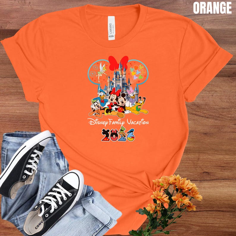 May include: Orange t-shirt featuring a Disney Family Vacation design with Mickey and Minnie Mouse, Donald Duck, and other characters. The design includes a castle and the year 2026. The shirt is paired with blue jeans and black sneakers.