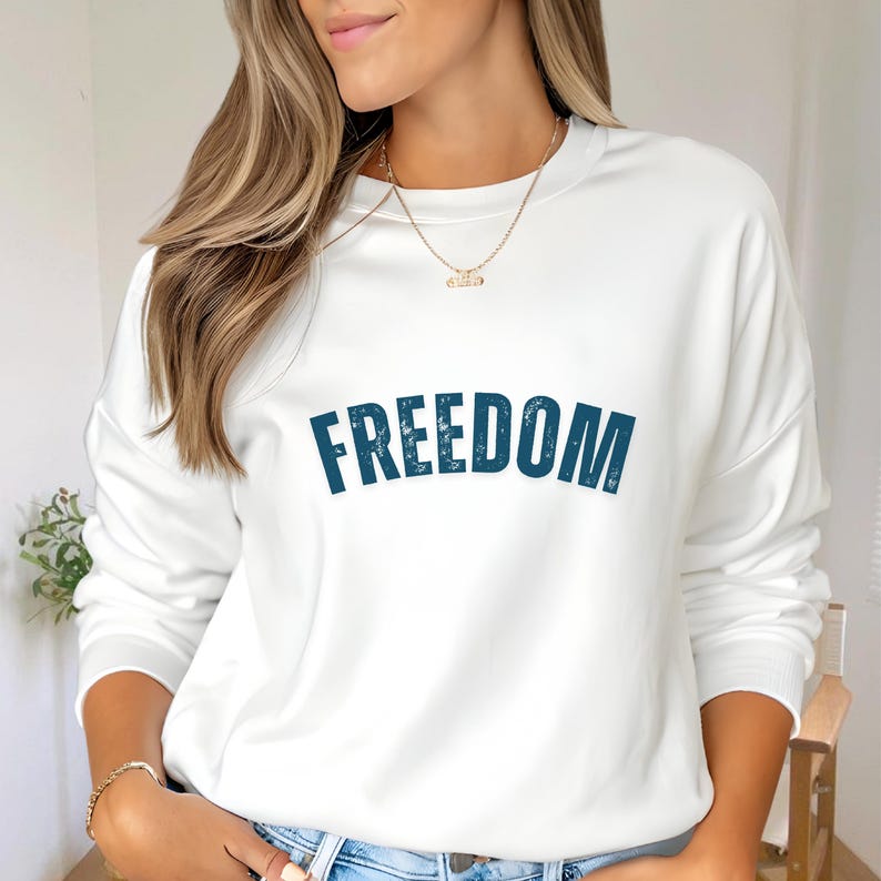 Puede incluir: Sudadera blanca con la palabra "FREEDOM" impresa en una fuente desgastada de color verde azulado oscuro en el pecho. La sudadera tiene cuello redondo y mangas largas. El estilo es informal y c&oacute;modo.