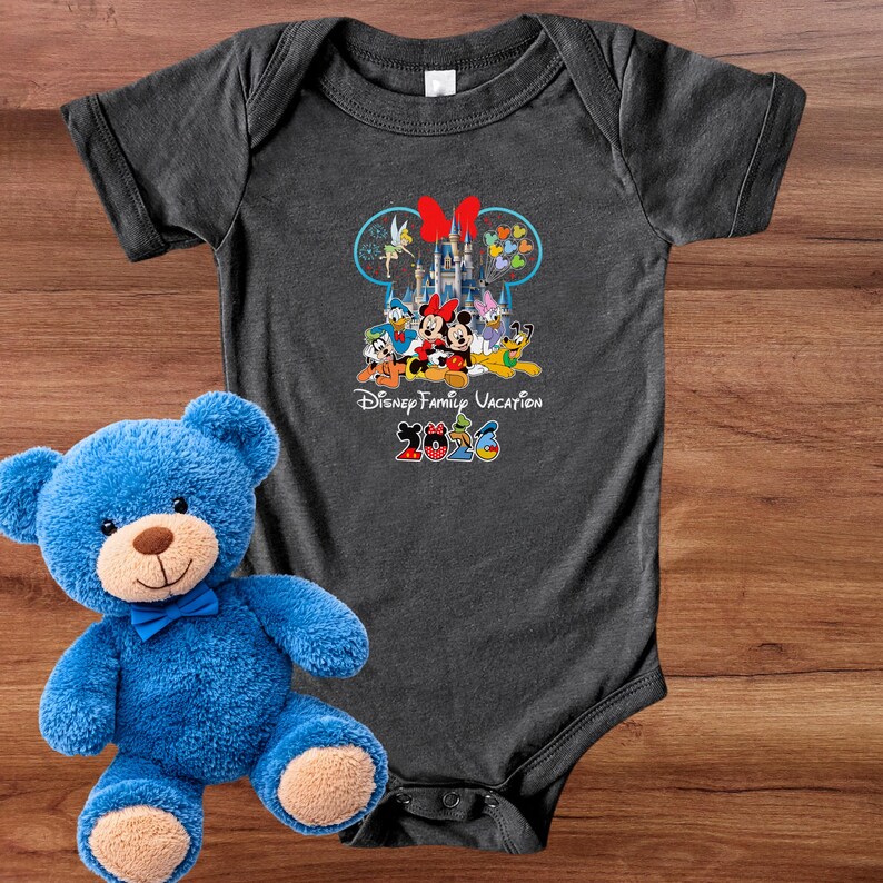 May include: Dark gray baby bodysuit featuring a colorful graphic of Disney characters, a castle, and the text "Disney Family Vacation 2023". A blue teddy bear with a blue bow tie is next to the bodysuit.